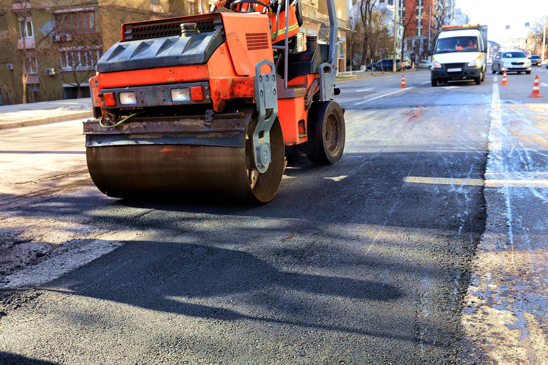 A heavy vibratory roller compacts hot asphalt on a repaired asphalt surface in the middle of the roadway on a clear sunny day.